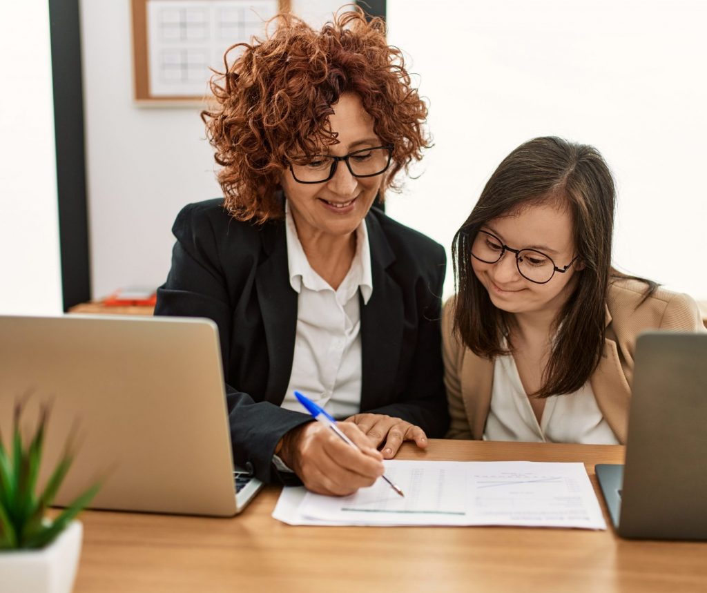 Group of two women working at the office. Mature woman and down syndrome girl working at inclusive teamwork. transition to adulthood